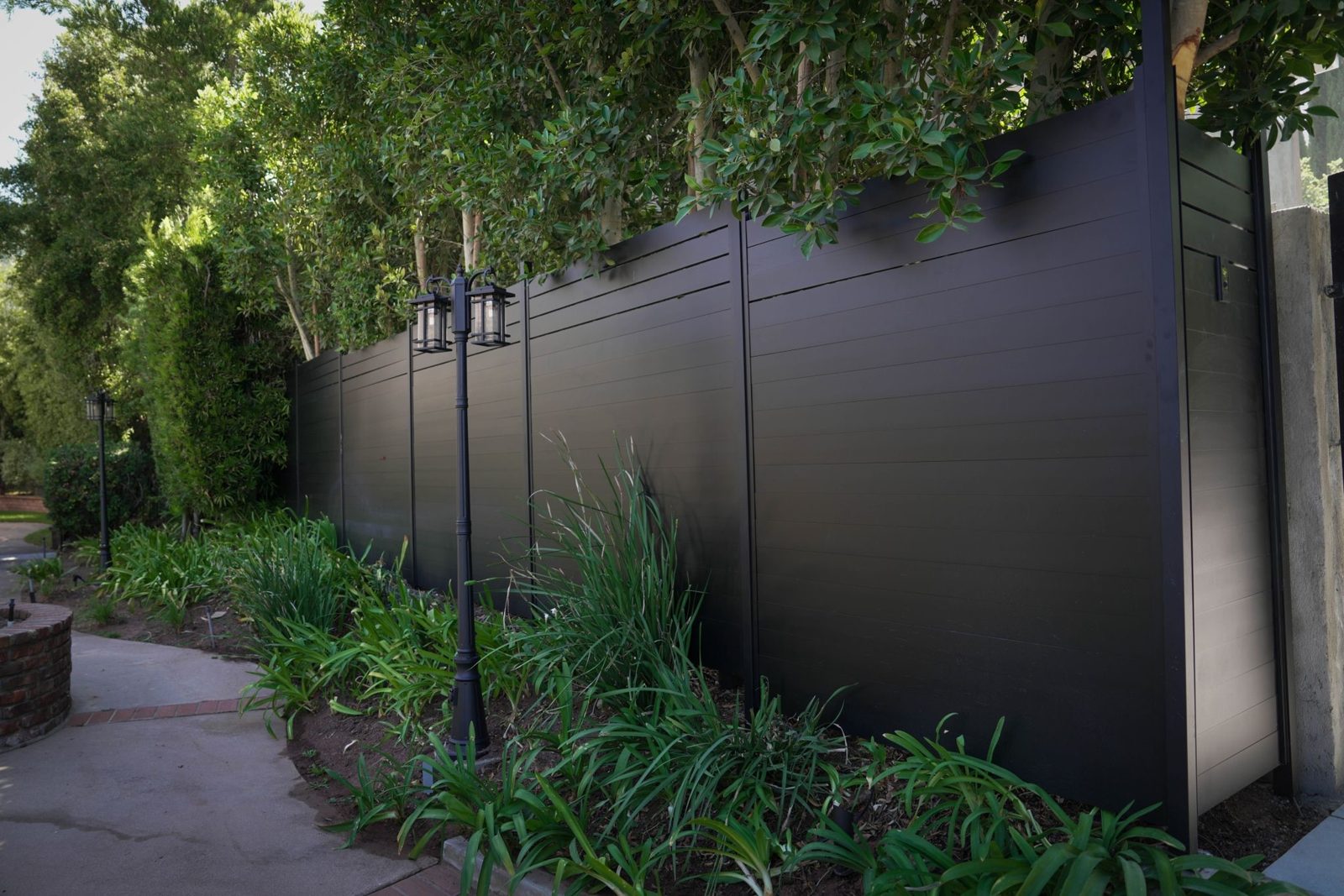 Tall black privacy fence along a garden path with lamp posts